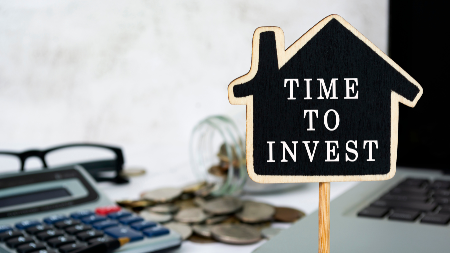 A small wooden sign in the shape of a house with the words "TIME TO INVEST" placed next to coins, a calculator, and a laptop.