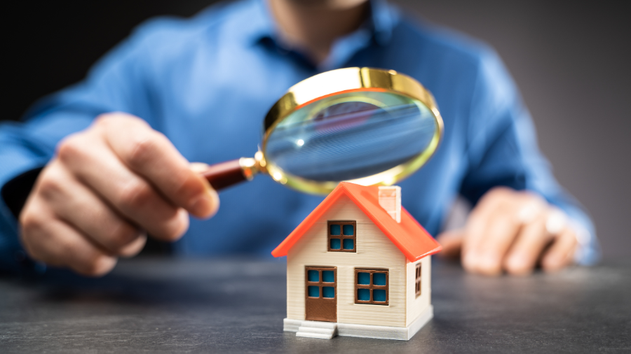 Person holding a magnifying glass examining a small model house, symbolizing home inspection or property investment.