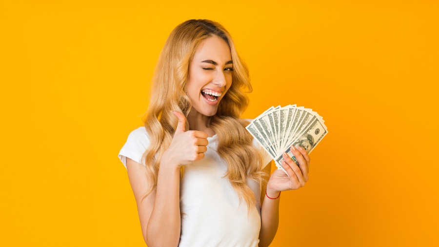 A smiling woman winking, giving a thumbs up, and holding a fan of hundred-dollar bills against a bright yellow background.