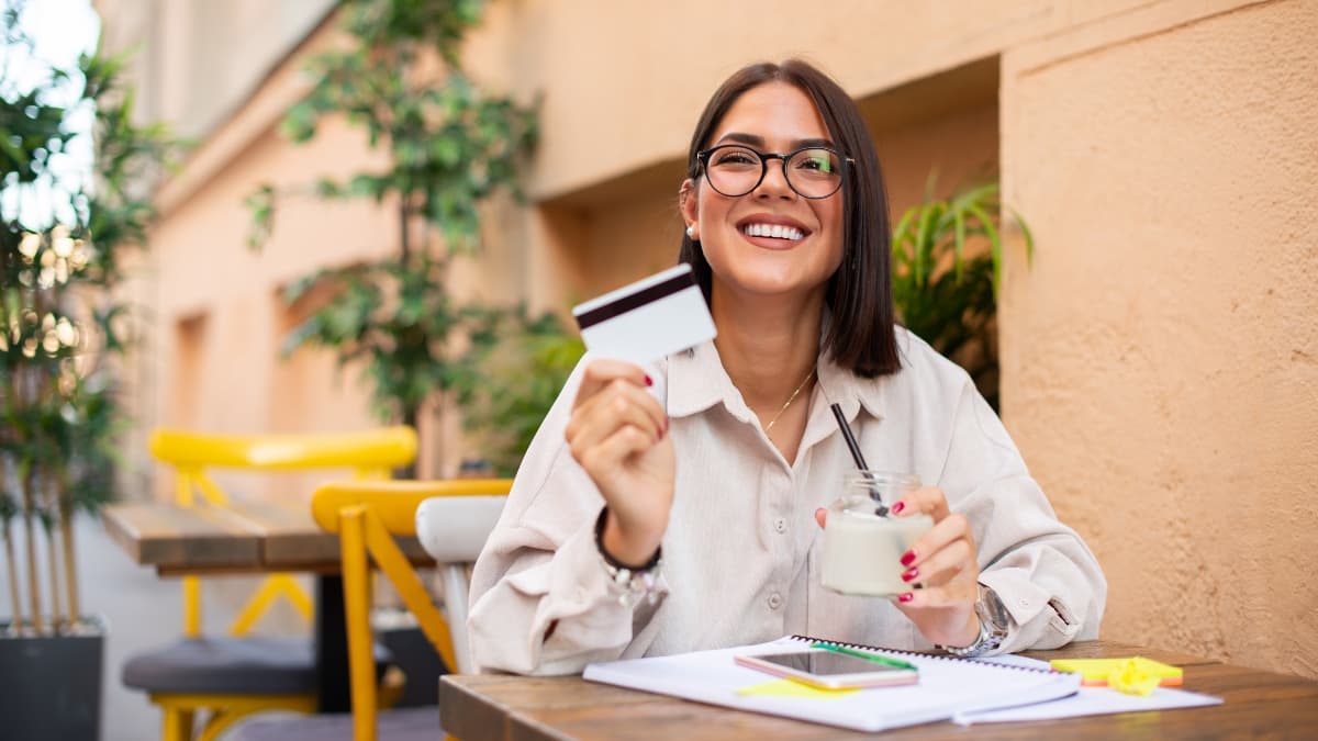 Smiling woman holding a credit card and yogurt jar while sitting at an outdoor café table.
