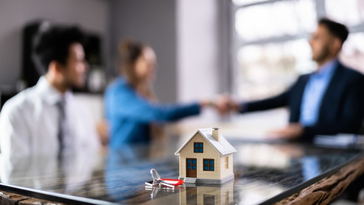 A small model house with keys on a table while two people shake hands in the background during a home loan meeting.