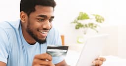 Smiling man holding a credit card while looking at a tablet at home, representing online credit card account review and financial planning